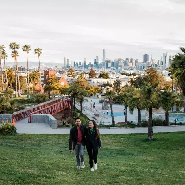 Um casal caminha em direção à câmera com Dolores Park e o horizonte de 贝博体彩app atrás deles.